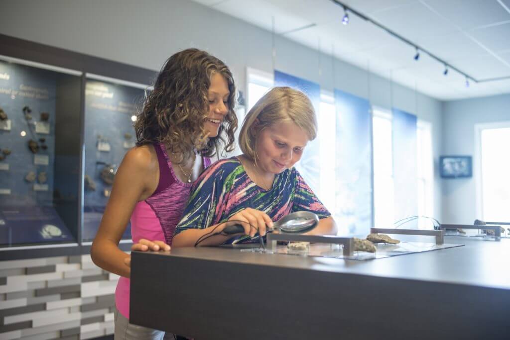 Deux jeunes filles observent des fossiles à la loupe dans l’exposition au centre d’accueil de Havre-Saint-Pierre. Réserve de parc national de l’Archipel-de-Mingan / Two young girls looking through a magnifying glass at fossils on exhibit at the Havre-Saint-Pierre reception and interpretation centre. Mingan Archipelago National Park Reserve